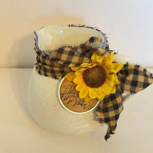 May include: A speckled white ceramic vase with a black and tan plaid ribbon and a yellow sunflower. A small, round, metallic tag with illegible text is attached to the vase.