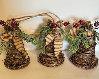 May include: Three brown twig bells with red berries, pine sprigs, and a tan and red striped ribbon. The bells are strung together with twine.