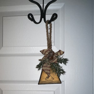 May include: A rustic, triangular-shaped bell with a burlap bow and pine sprigs hanging from a black hook. The bell is decorated with a vintage-style image and has a brown, wooden frame.