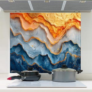 Op de afbeelding: Een keukenachterwand met een abstract ontwerp in blauw, goud en oranje tinten. Het kunstwerk lijkt op gelaagde rotsformaties. Daaronder staan op een witte kookplaat een zwarte koekenpan, een kleine zwarte pot en een grijze pot.