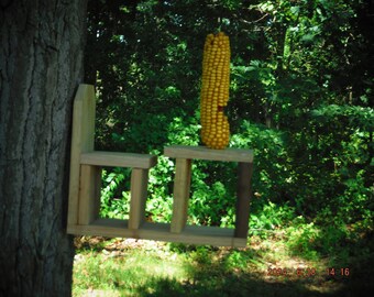 May include: A wooden bird feeder with three shelves, attached to a tree trunk. A single ear of corn hangs from the top shelf.