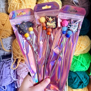May include: Three sets of knitting needle sets in iridescent pink packaging. Each set features a different character charm, including a bear, an Eevee, and a bunny. The needles have colorful bead accents and are displayed in front of various yarn skeins.