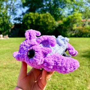 May include: A plush toy shaped like an axolotl, crafted from soft, textured yarn in shades of bright purple and light blue. The creature features large, round, black plastic eyes and prominent, feathery external gills on its head.