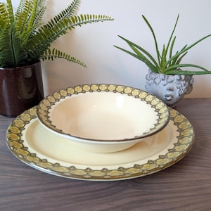 Puede incluir: Un juego de cuenco y plato de color crema con un borde decorativo dorado y verde oliva. El cuenco se encuentra encima del plato, y ambos están sobre una superficie de madera. El juego forma parte de una colección de vajilla vintage.