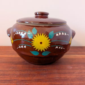 Può includere: Un vaso di ceramica marrone con coperchio, decorato con fiori gialli e foglie verdi. Il vaso ha una smaltatura bianca ed è dipinto a mano.