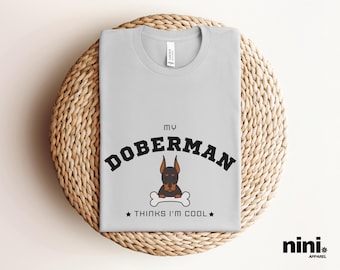 Op de afbeelding: Een lichtgrijs T-shirt met de tekst "My Doberman thinks I'm cool" in zwarte letters. Een cartoon Dobermann met bruin en zwart haar bevindt zich boven de tekst met een bot in zijn bek.