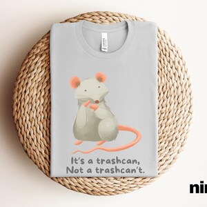 Peut inclure: Un t-shirt gris clair avec une illustration de souris grise de dessin animé avec des oreilles et un nez roses. La souris tient un morceau de fromage dans ses pattes. Le texte "It's a trashcan, Not a trashcan't." est imprimé sous la souris.