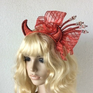 May include: A red headband featuring a glittery devil horn on one side and a large, sheer, red ribbon bow on the other. The bow is accented with four thin, red, glittery stems, each topped with a small, white, detailed skull figurine.