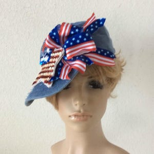 May include: A denim baseball cap with a decorative bow featuring red, white, and blue ribbons and sequin accents. The bow is designed with American flag patterns, adding a patriotic touch to the hat.