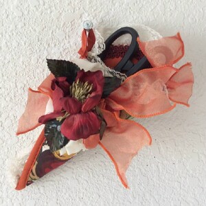 May include: A cone-shaped sewing needle holder with a red flower, orange ribbon, and black scissors. The holder is decorated with lace and a floral pattern.