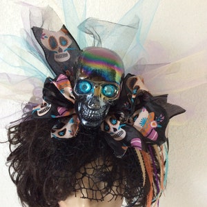 May include: A black skull with iridescent paint and blue eyes is attached to a black and white ribbon with a sugar skull pattern. The ribbon is tied into a bow and is surrounded by layers of tulle in shades of blue, purple, and white. The skull is worn on a black wig with a black lace veil.