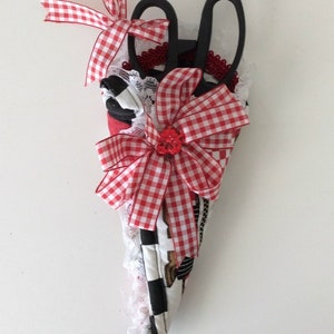 May include: A red and white checkered fabric scissor holder with a white lace trim and a red bow with a red button in the center. The holder is filled with two black scissors.