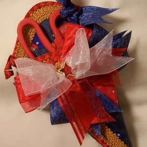 May include: A red, white, and blue fabric cone with gold trim, decorated with a red, white, and blue ribbon bow and a pair of red scissors.