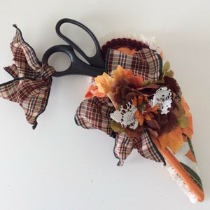 May include: A fabric scissor holder with a plaid bow, fall leaves, and a flower. The holder is shaped like a cone and has a lace trim.