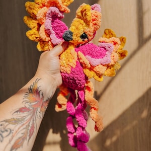 May include: A crocheted phoenix plush toy featuring vibrant layers of yarn in shades of bright orange, yellow, and hot pink. The bird has a small black beak, dark plastic eyes, and long, curly tail feathers that extend downward from the body.