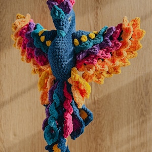 May include: A vibrant, handmade crochet bird with outstretched wings. The bird features a blue body, a multi-colored head, and wings with shades of orange, pink, purple, and teal. Yellow accents are scattered throughout the design. The bird is displayed against a wooden background.