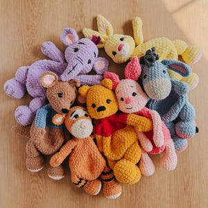 May include: A collection of seven crocheted plush toys featuring characters including a yellow bear in a red shirt, a pink piglet, a blue donkey, a brown kangaroo, an orange tiger, a purple elephant, and a yellow rabbit, all made of soft yarn.