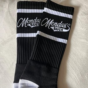 May include: Pair of black socks with white stripes at the top and the text "Monday's Suck" in white script. The socks have a white heel and toe, and a ribbed design.