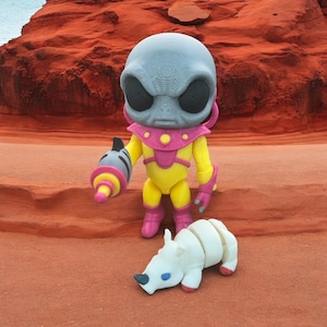 May include: A gray alien figurine with a yellow and pink spacesuit holding a pink and yellow ray gun. The alien is standing on a red surface with a white rhinoceros figurine lying on its side.