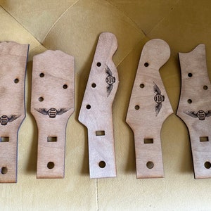 Puede incluir: Cinco palas de guitarra de madera sin terminar de varias formas. Cada pala tiene agujeros pre-perforados para las clavijas y un logotipo grabado con láser con un emblema alado. La madera es de color claro y natural.