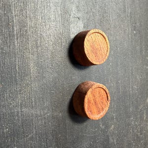 May include: Two small, round wooden objects with a reddish-brown hue. Each has a circular, recessed design on the top surface. The objects are set against a dark, textured background.