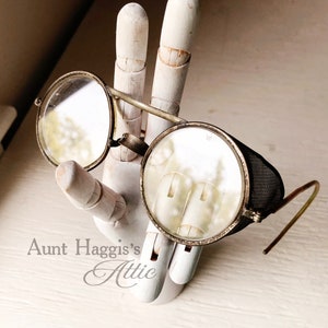 May include: Antique round eyeglasses with metal frames and clear lenses. One lens has a mesh side shield. The glasses are displayed on a white wooden hand. The text "Aunt Haggis's Attic" is visible.
