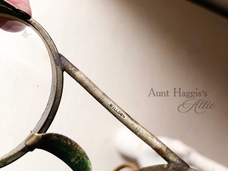 May include: Close-up of antique Willson eyeglasses with a silver-toned metal frame. The glasses have round lenses and a bridge. The text "WILLSON" is visible on the arm. The image is from Aunt Haggis's Attic.
