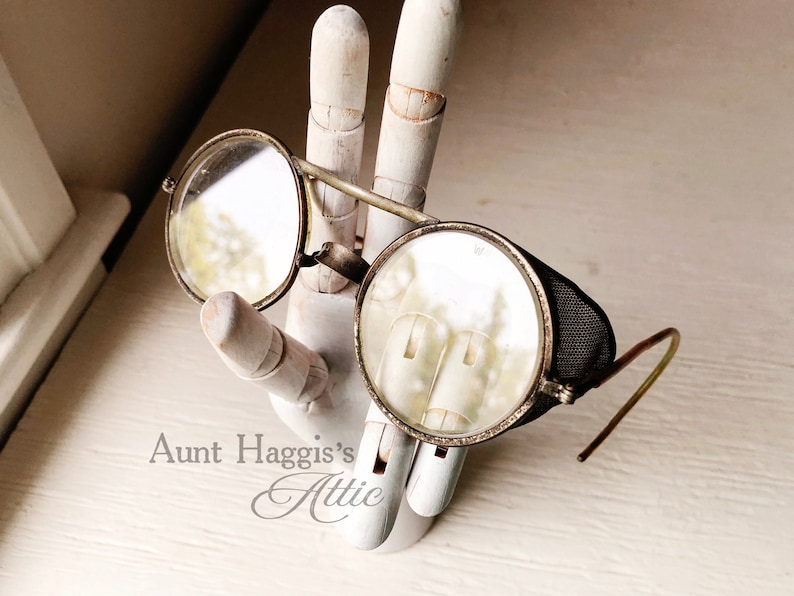 May include: Antique round eyeglasses with metal frames and clear lenses. One lens has a mesh side shield. The glasses are displayed on a white wooden hand. The text "Aunt Haggis's Attic" is visible.