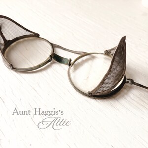 May include: Antique steampunk-style eyeglasses with round lenses and mesh side shields. The glasses have a metal frame with a dark finish and a wooden arm. The design is reminiscent of vintage industrial or laboratory eyewear.