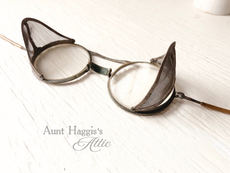 May include: Antique steampunk-style eyeglasses with round lenses and mesh side shields. The glasses have a metal frame with a dark finish and a wooden arm. The design is reminiscent of vintage industrial or laboratory eyewear.