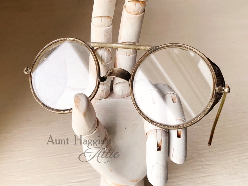 May include: Antique round eyeglasses with a silver-colored frame and clear lenses. The glasses have small side shields and thin, curved temples. The image includes the text "Aunt Haggis's Attic".