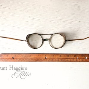 May include: Antique round eyeglasses with wire frames and curved temples. The glasses have clear lenses and small side shields. A wooden ruler is placed below the glasses. The text "Aunt Haggis's Attic" is at the bottom.