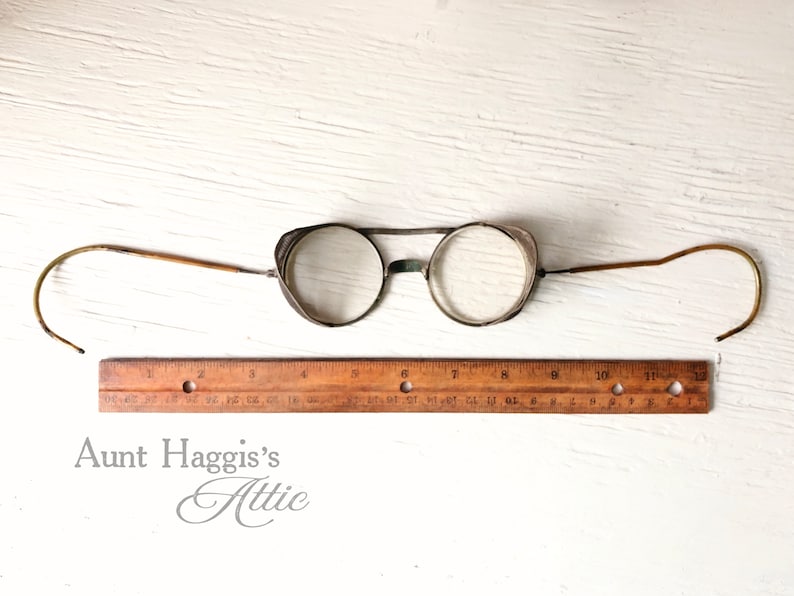 May include: Antique round eyeglasses with wire frames and curved temples. The glasses have clear lenses and small side shields. A wooden ruler is placed below the glasses. The text "Aunt Haggis's Attic" is at the bottom.