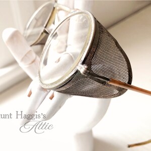 May include: Vintage steampunk-style goggles with clear lenses and a mesh side shield. The goggles have a metal frame and brown temples. The image is set against a white background.