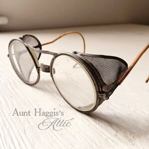 May include: Antique metal safety glasses with clear lenses and mesh side shields. The glasses have a vintage aesthetic, with a weathered appearance and curved temples. The text "Aunt Haggis's Attic" is visible.