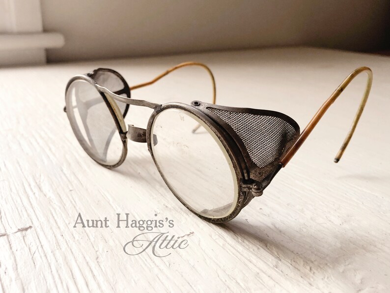 May include: Antique metal safety glasses with clear lenses and mesh side shields. The glasses have a vintage aesthetic, with a weathered appearance and curved temples. The text "Aunt Haggis's Attic" is visible.