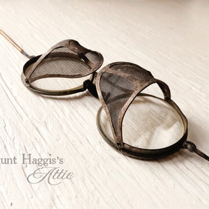May include: Antique steampunk-style goggles with mesh eye covers and clear lenses. The goggles have a dark metal frame and slender arms. The text "Aunt Haggis's Attic" is visible in the lower left corner.