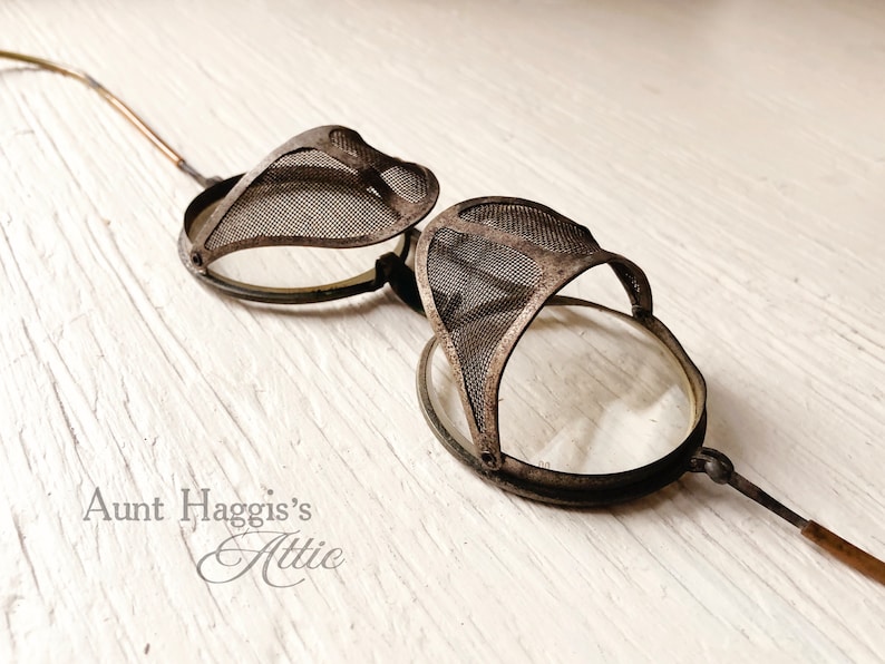 May include: Antique steampunk-style goggles with mesh eye covers and clear lenses. The goggles have a dark metal frame and slender arms. The text "Aunt Haggis's Attic" is visible in the lower left corner.