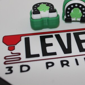 Può includere: Due tasti per tastiera stampati in 3D, verdi e bianchi, con un trifoglio e un ferro di cavallo. I tasti per tastiera sono su una superficie bianca con il testo "LEVEL UP 3D PRINTING".