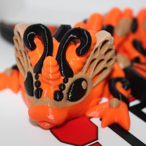 Peut inclure: Figurines de dragon orange et noir imprimées en 3D avec des ailes et une corne. Le dragon a un design détaillé avec des accents noirs et un masque à motifs marrons.
