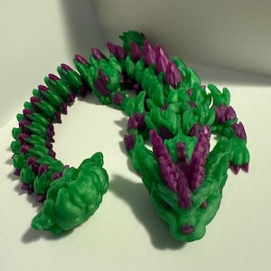 Peut inclure: Une figurine de dragon imprimée en 3D de couleur verte et violette avec un dos hérissé et une queue enroulée.