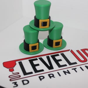 Può includere: Tre cappelli da folletto verdi e neri impilati uno sopra l'altro. I cappelli hanno una fibbia dorata e sono realizzati in plastica stampata in 3D.