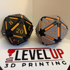 Puede incluir: Dos dados D20 grandes impresos en 3D. Uno es negro con números amarillos, el otro es gris con números naranjas. Los dados están sobre una superficie blanca con el texto "LEVEL UP 3D PRINTING" en negro y rojo.