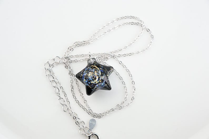 May include: A silver chain necklace with a black star-shaped pendant. The pendant has a swirling design of gold and blue glitter.