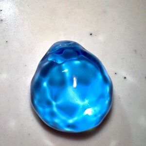 Puede incluir: Una esfera de vidrio azul brillante con una superficie lisa y redondeada. La esfera refleja la luz, creando un efecto brillante.