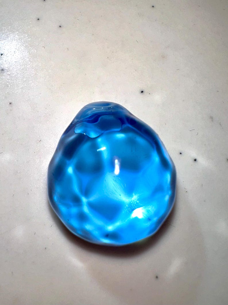 Puede incluir: Una esfera de vidrio azul brillante con una superficie lisa y redondeada. La esfera refleja la luz, creando un efecto brillante.