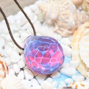Puede incluir: Un colgante de vidrio rosa y azul con un patrón de remolino, colgando de un cordón marrón. El colgante está rodeado de piedras blancas y conchas.