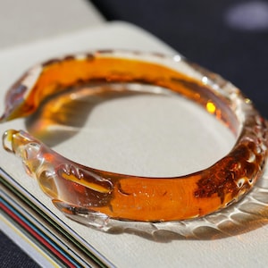May include: A clear amber glass bangle bracelet with a textured surface. The bracelet is shaped like a dragon with intricate details.