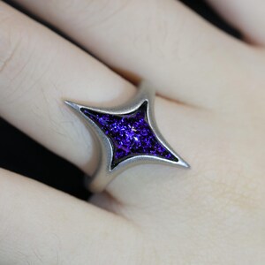 May include: A silver ring featuring a four-pointed star design. The star is filled with a sparkly purple resin.