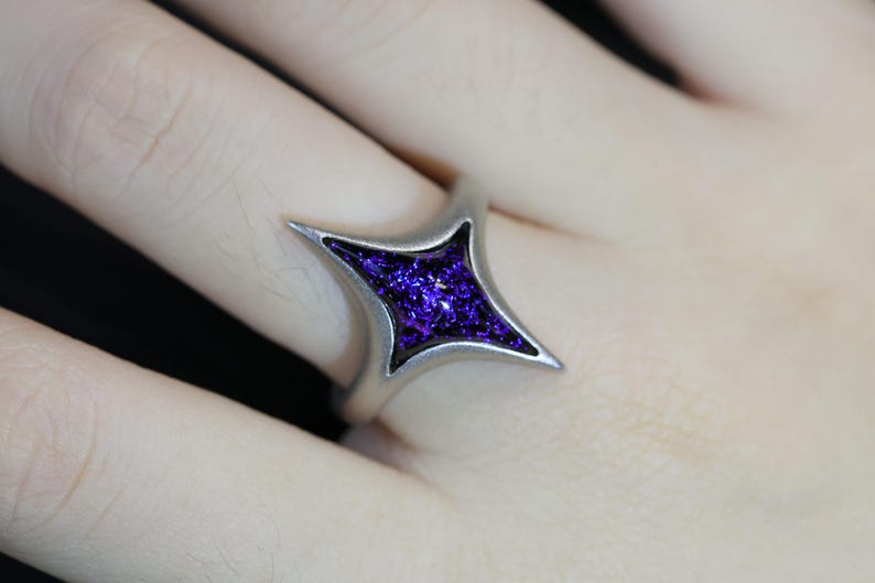 May include: A silver ring featuring a four-pointed star design. The star is filled with a sparkly purple resin.